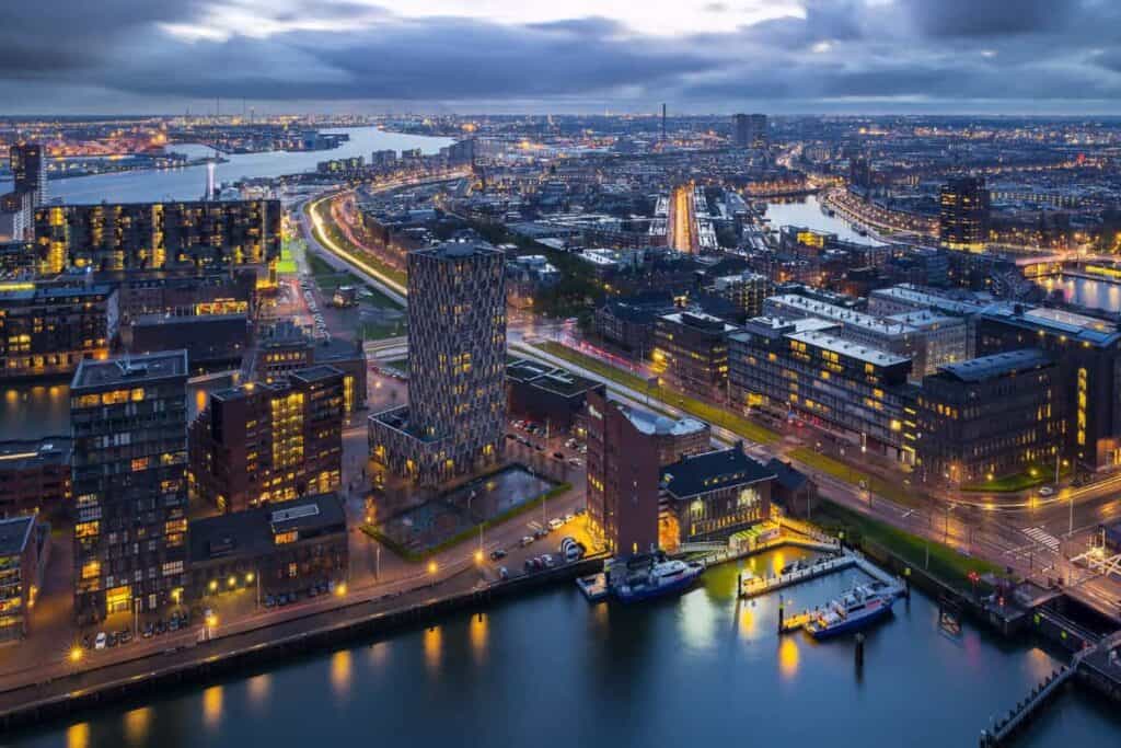 Stand de exposición Vista aérea de una ciudad moderna con edificios y calles iluminadas al anochecer, flanqueada por vías fluviales y puentes, con barcos atracados a lo largo del canal y un cielo nublado sobre sus cabezas.