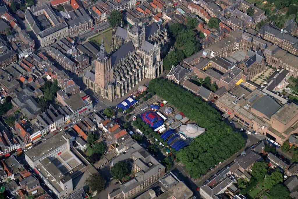 Stand de exposición Vista aérea de una gran catedral gótica rodeada de edificios de la ciudad, con un parque verde y coloridas carpas o estructuras de feria cercanas, en una ciudad europea densamente urbanizada.