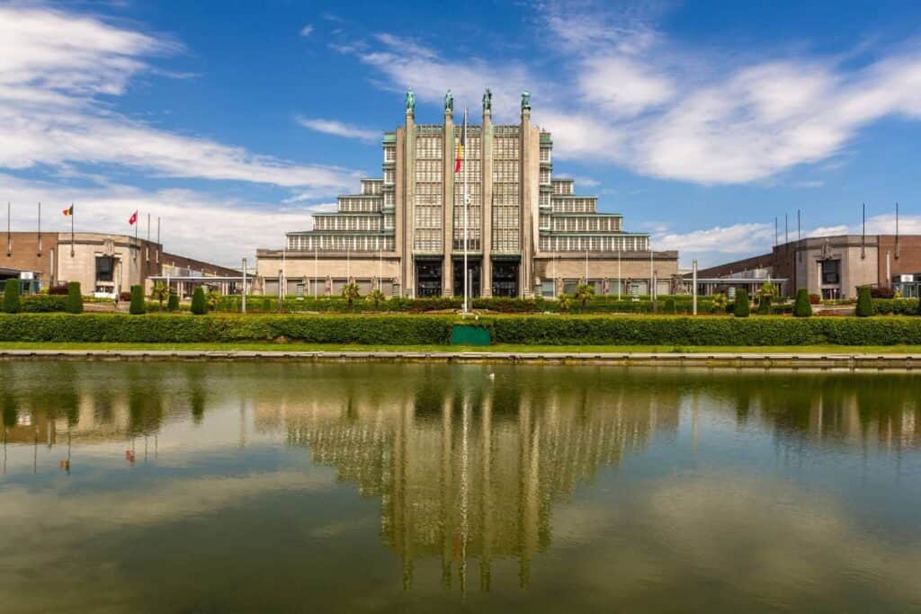 Beursstand Art-decogebouw met gelaagde, symmetrische architectuur, grote verticale zuilen, beelden bovenop en glazen ramen, weerspiegeld in een kalme rechthoekige vijver onder een blauwe hemel met verspreide wolken.