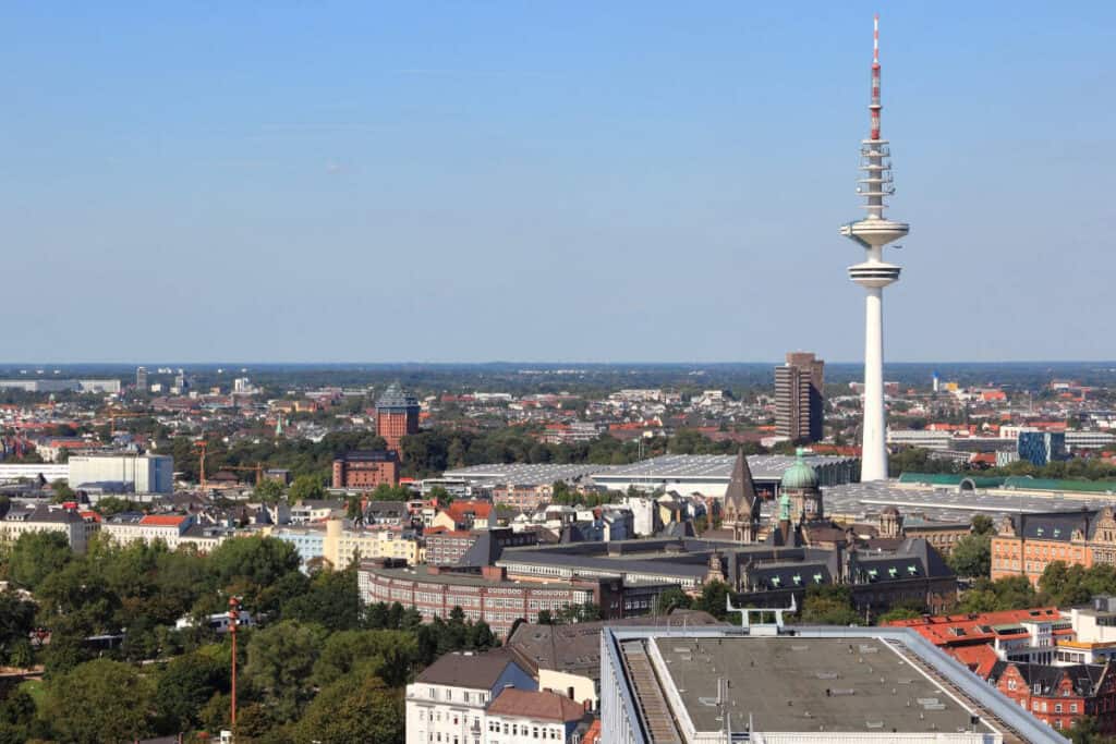 Beursstand Een stadsgezicht met een mix van historische en moderne gebouwen, waaronder een hoge witte communicatietoren op de achtergrond, omringd door groen en een helderblauwe lucht.