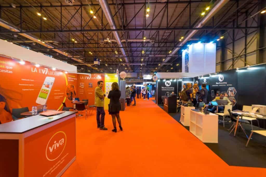 Beursstand Een helder verlichte tentoonstellingshal met stands aan beide kanten, mensen die over een oranje tapijt lopen en praten, en verschillende displays, spandoeken en technologie die door de hele ruimte te zien zijn.