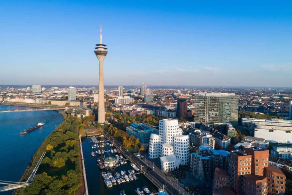 Beursstand Luchtfoto van de skyline van Düsseldorf met de Rheinturm toren, moderne gebouwen bij MedienHafen, de rivier de Rijn, groene ruimtes en boten aangemeerd in een jachthaven onder een strakblauwe hemel.