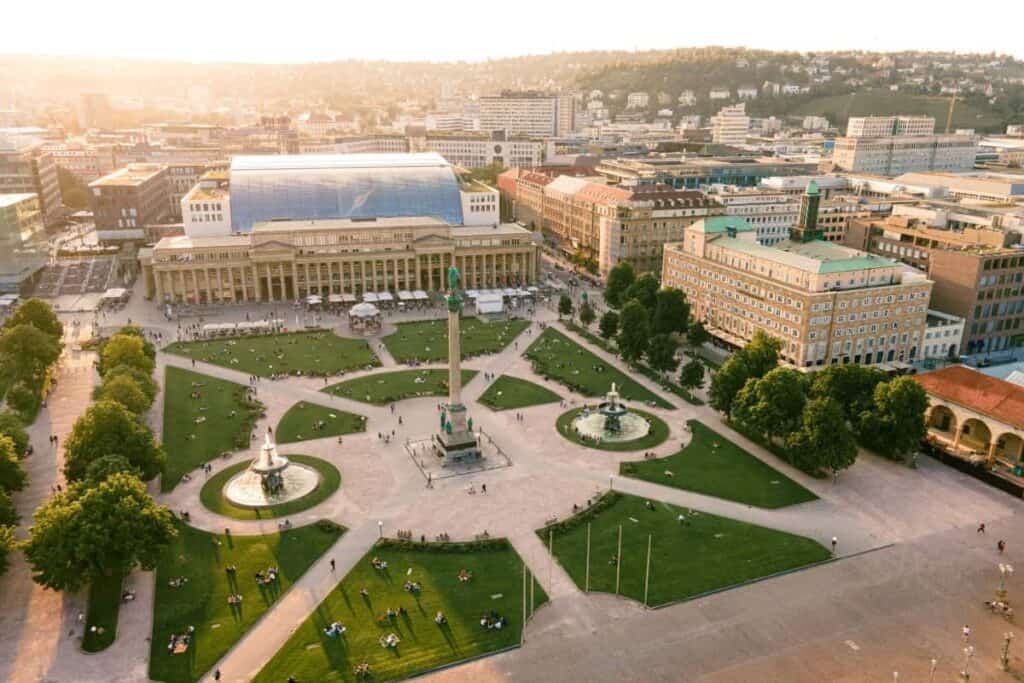Beursstand Luchtfoto van Schlossplatz in Stuttgart, Duitsland, met groene gazons, fonteinen, ontspannende mensen en historische gebouwen rond het plein op een zonnige dag.