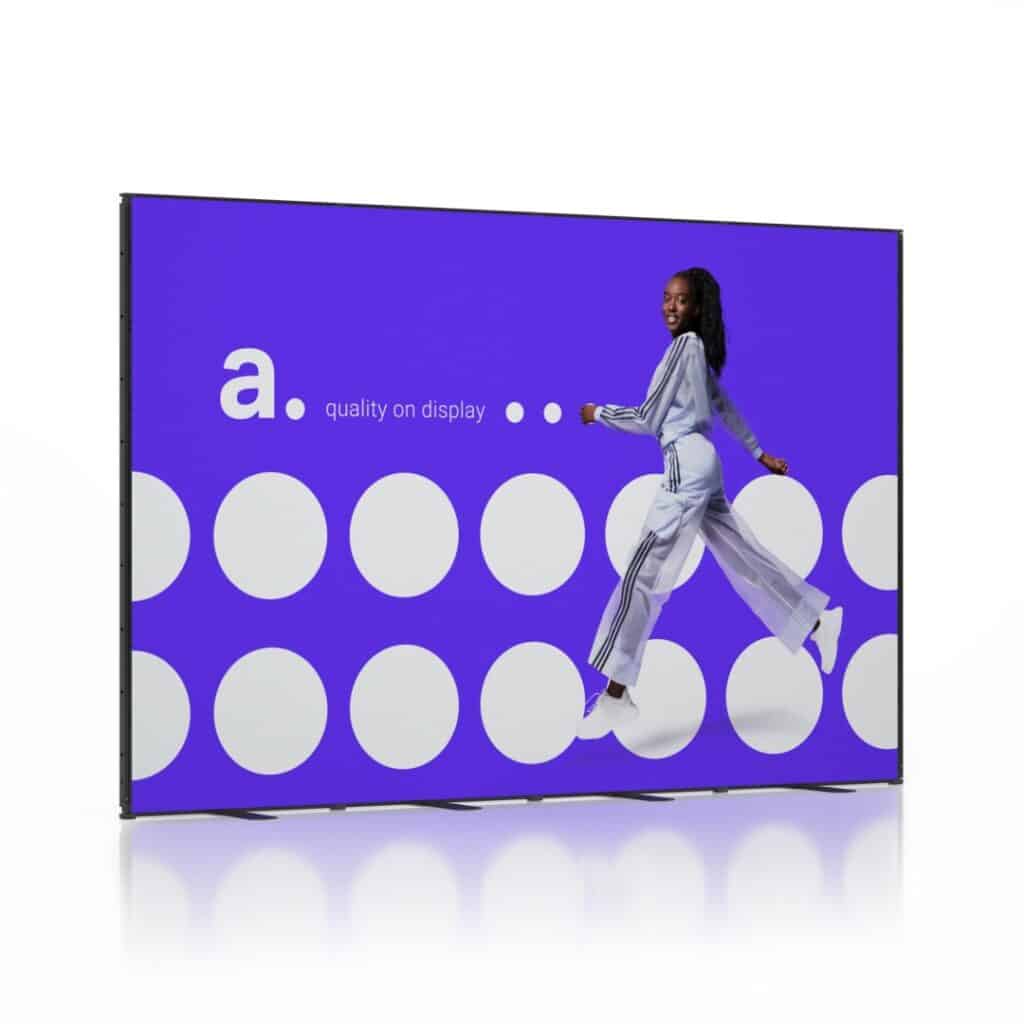 Beursstand Op een groot digitaal scherm, de Multiframe 4 x 3 meter - Kopopstelling, is een vrouw in trainingspak te zien die springt voor een fel paarse achtergrond met witte stippen, de letter "a." en de zin "quality on display".