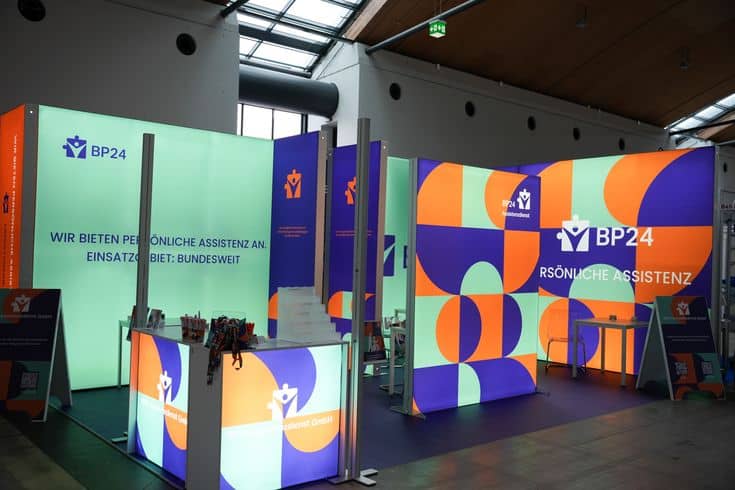 Stand d’exposition Un stand d’exposition vibrant et moderne pour BP24 présente des motifs géométriques colorés, des panneaux illuminés et une signalétique en allemand offrant une assistance personnalisée. Des bureaux, des chaises et des objets promotionnels sont visibles dans l’installation.
