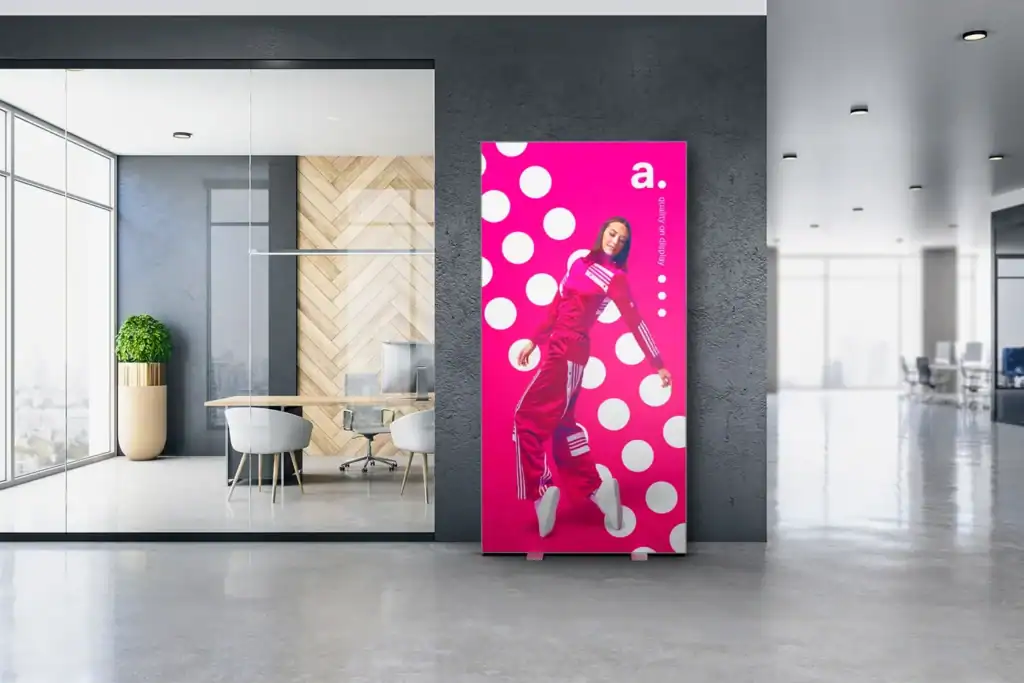 Ausstellungsstand Ein knallpinkes Poster mit großen weißen Punkten zeigt eine Frau in einem roten Overall, die eine Pose einnimmt. Es befindet sich in einer modernen Bürolobby in der Nähe eines Besprechungsraums mit Glaswänden, Holzakzenten und minimalistischem Dekor.