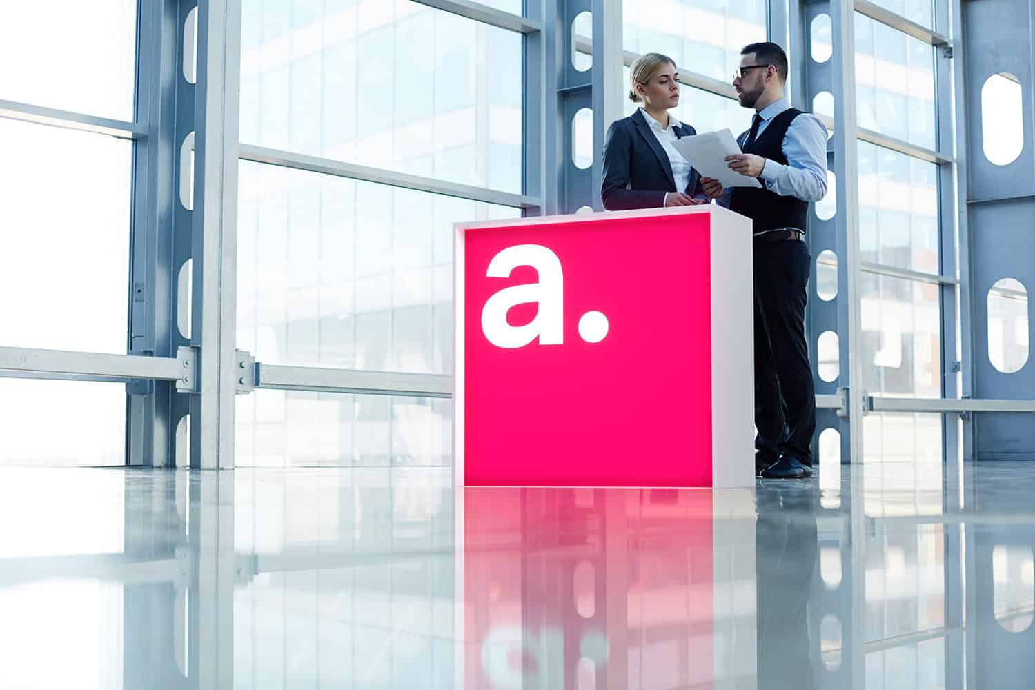 Beursstand Twee mensen in zakelijke kleding staan achter de Portable LED balie - Expo 100x100cm, een felroze receptiebalie met een grote witte letter "a", documenten te bespreken in een modern kantoor met glazen wanden.