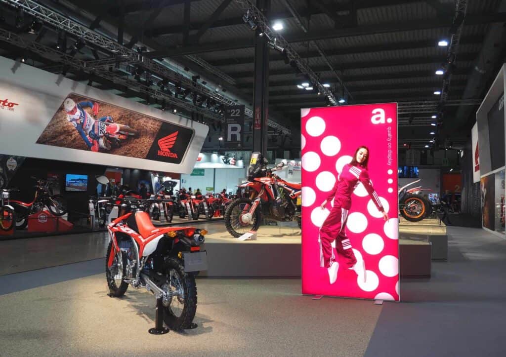 Stand d’exposition de motos Honda exposées dans un showroom ; un grand panneau rose avec une femme en combinaison de course et des points blancs se trouve à droite, tandis qu’une bannière Honda motocross est accrochée au mur de gauche au-dessus des motos.